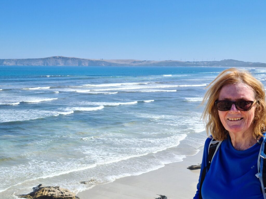 A close-up of a woman with blonde hair and sunglasses smiling at the camera. She is positioned on the right side of the frame, wearing a blue hiking shirt. In the background, a vast coastline stretches out with rolling blue waves breaking on a sandy beach under a clear sky. A distant headland with wind turbines is visible on the horizon.