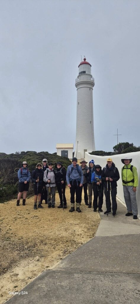 Group of twelve hikers in outdoor gear standing in front of a tall white lighthouse with a red top; adjacent small building and sandy terrain visible; concrete path leads to the lighthouse; cloudy sky overhead with vegetation in the background.