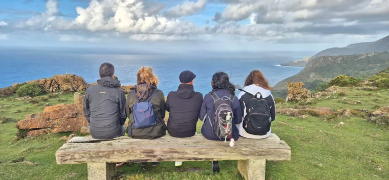 Five hikers with backpacks sitting on a rustic wooden bench, facing the ocean and rugged cliffs under a partly cloudy sky; grassy and rocky terrain in the foreground, with distant hills along the coastline.