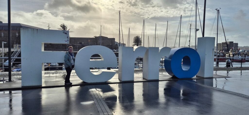 Large 3D sign spelling “Ferrol” at a waterfront marina, with a person standing, sailboats, palm trees, and buildings visible in the background under a cloudy sky with wet ground from recent rain.