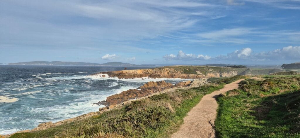 Coastal landscape with a dirt path winding through grassy terrain toward rocky cliffs where waves crash against the shore; deep blue sea with white surf, distant hills, and a partly cloudy sky.