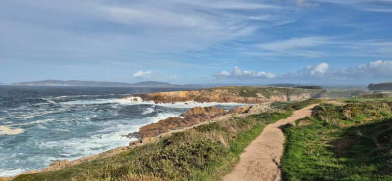 Coastal landscape with a dirt path winding through grassy terrain toward rocky cliffs where waves crash against the shore; deep blue sea with white surf, distant hills, and a partly cloudy sky.