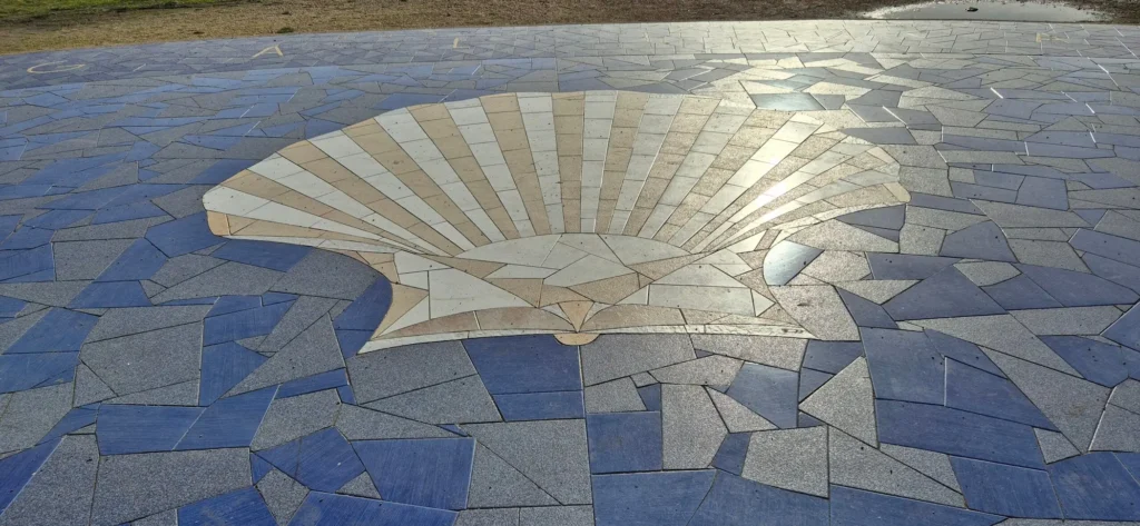 Large outdoor mosaic embedded in the ground, featuring a light‑colored scallop shell design at the center with tiles radiating outward, surrounded by irregular blue and gray tiles; sandy terrain visible nearby.