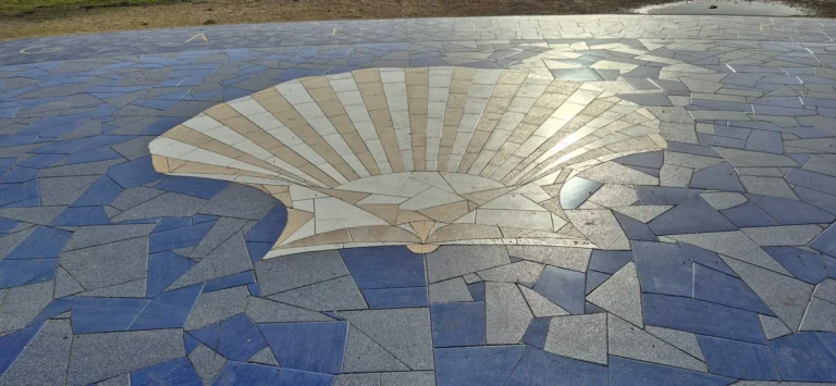 Large outdoor mosaic embedded in the ground, featuring a light‑colored scallop shell design at the center with tiles radiating outward, surrounded by irregular blue and gray tiles; sandy terrain visible nearby.