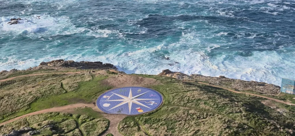 Scenic coastal viewpoint with a large circular mosaic compass rose in vivid blue embedded in grassy terrain near a cliff edge; ocean waves crash against rocky shoreline in the background under a partly cloudy sky.