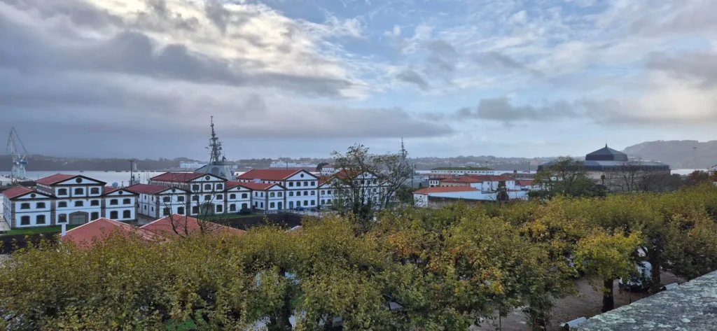 Scenic coastal town with white buildings featuring red-tiled roofs and black trim, arranged in a grid near the waterfront; a large ship and cranes visible in the background, autumn trees in the foreground, and hills under a partly cloudy sky.
