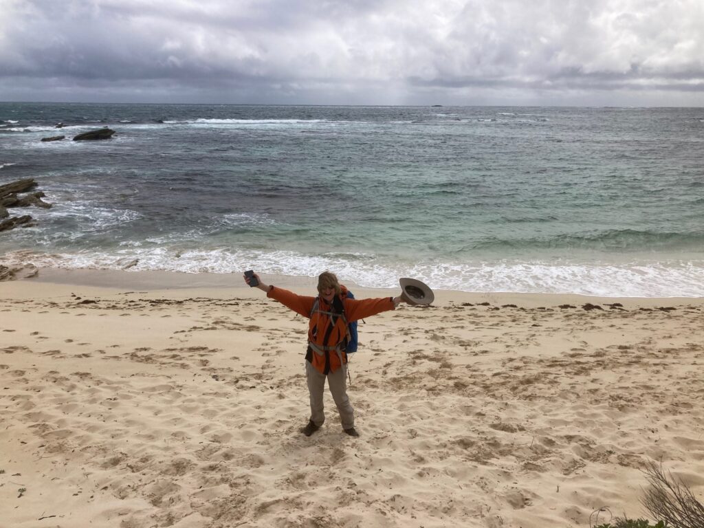 A hiker stands on a wide sandy beach with arms outstretched in celebration, holding a hat and phone, with the ocean and a cloudy sky in the background.