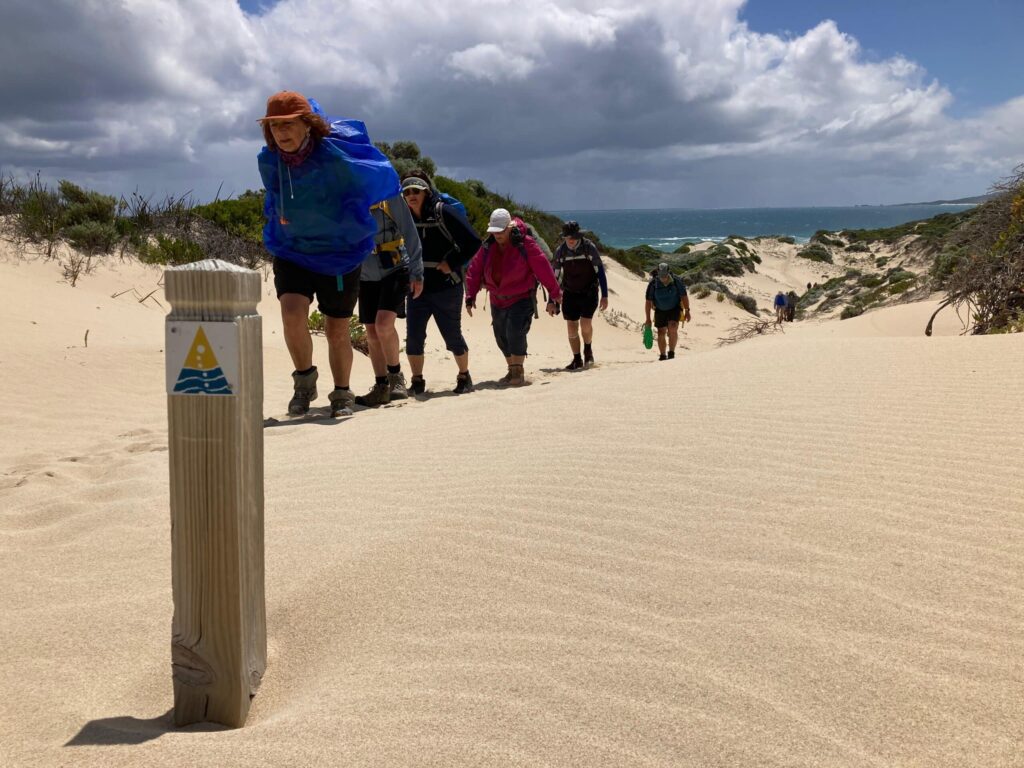 A line of hikers with backpacks climbs a tall, rippled sand dune toward a trail marker under a cloudy sky.