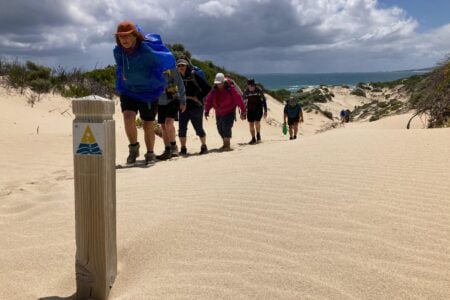 A line of hikers with backpacks climbs a tall, rippled sand dune toward a trail marker under a cloudy sky.