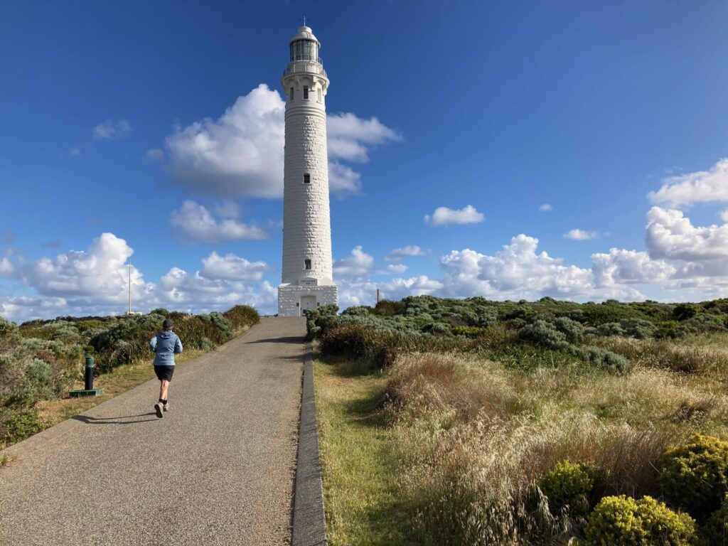 A person jogs up a paved path toward the tall, white limestone Cape Leeuwin Lighthouse under a blue sky with fluffy white clouds.