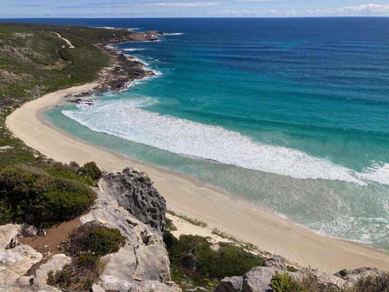 A high-angle view looking down at a long, crescent-shaped white sand beach with turquoise waves breaking on the shore, flanked by rocky cliffs and a winding coastal path.