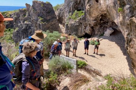 A group of hikers waving from a sandy area in front of a large, rugged sea cave along a rocky coastline.