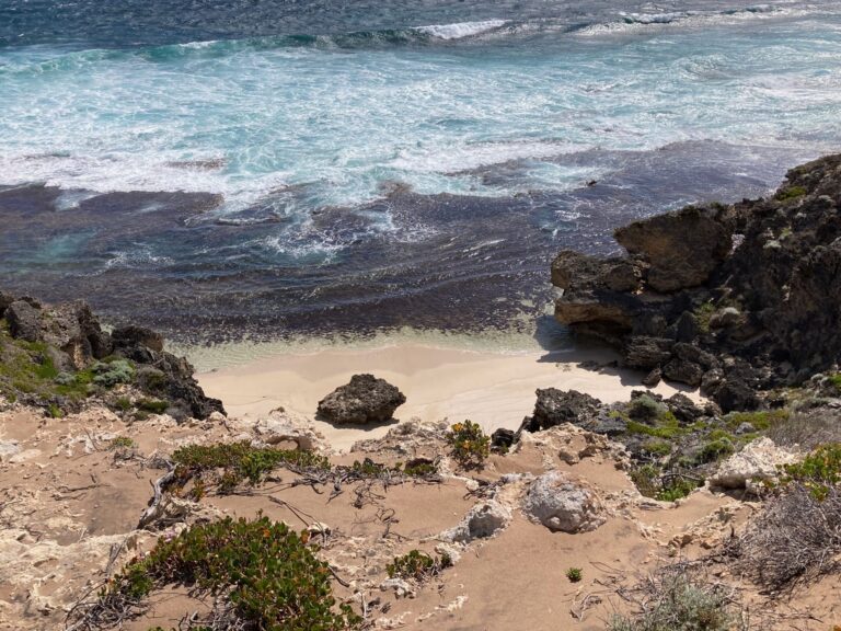 A high-angle view looking down at a small, secluded white sand beach nestled between rugged limestone cliffs and crashing turquoise waves.