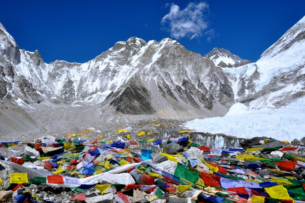 A wide view of Mount Everest Base Camp featuring colorful Buddhist prayer flags in the foreground against a backdrop of massive, snow-capped Himalayan peaks and a clear blue sky.