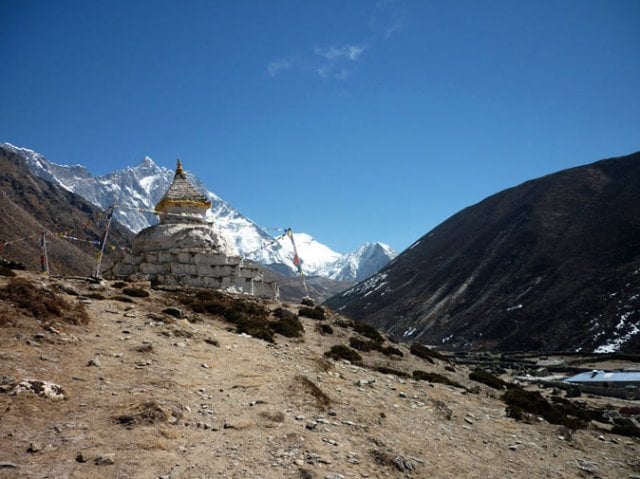 A small settlement of stone buildings with bright blue roofs nestled in a snowy, rocky valley at the foot of massive, snow-covered mountain peaks under a cloudy sky.