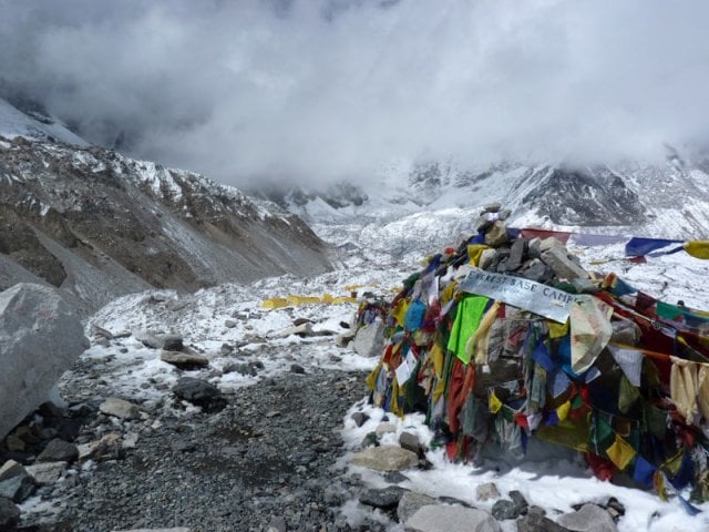 A remote outpost or lodge with several blue-roofed buildings sits on a snowy, rocky plain. In the background, a sharp, white mountain peak towers into a blue sky, partially draped in thick white clouds. Footprints and narrow paths crisscross the snow in the foreground.