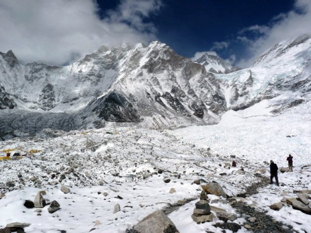 A cluster of simple stone buildings with vibrant blue roofs sits in a snow-dusted, rocky valley. In the background, a massive, jagged mountain peak is partially obscured by swirling white clouds under a blue sky.