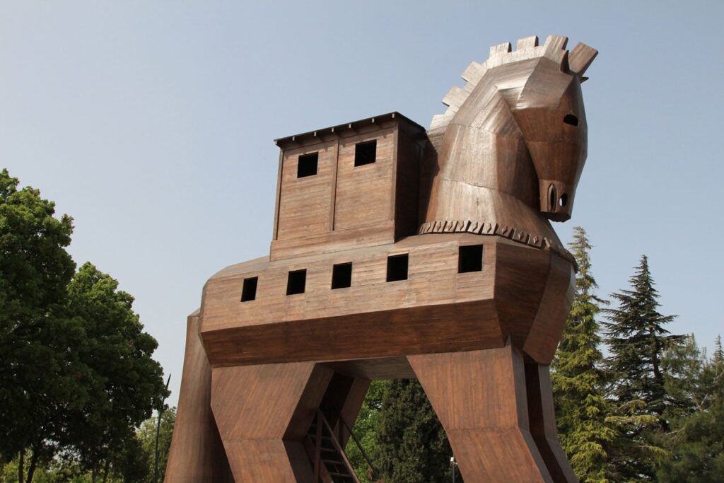 A large, multi-story wooden sculpture of a horse, featuring small windows and a built-in room on its back, set against a clear sky and green trees.