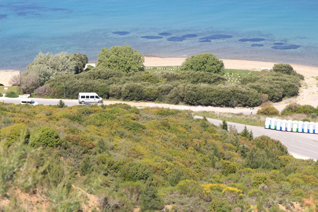 A high-angle landscape shot from a lush green hillside looking down toward a paved coastal road. A white van and several portable toilets are visible on the roadside. Beyond the road, dense green shrubbery borders a sandy beach and the clear, turquoise waters of a bay.