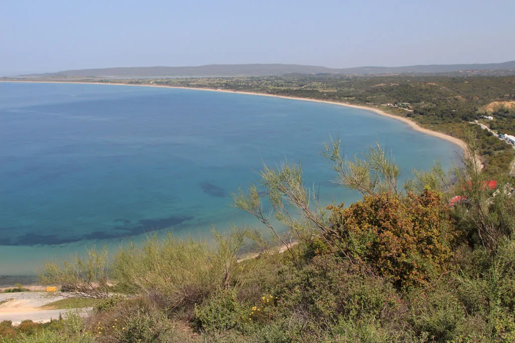 A panoramic high-angle view from a brush-covered hillside overlooking a wide, crescent-shaped bay. The calm blue water transitions to sandy shores, with rolling green hills and a hazy coastline visible in the far distance under a clear sky.