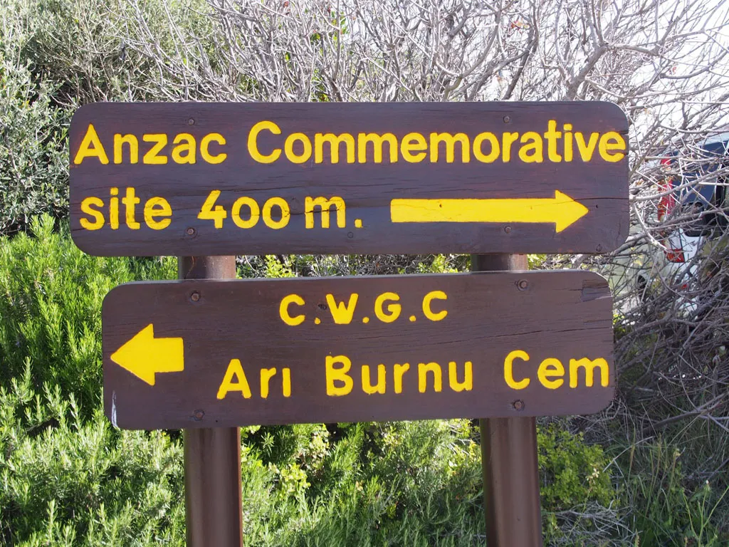 A close-up of two dark wooden directional signs with bright yellow lettering mounted on brown metal poles. The top sign points right with the text "Anzac Commemorative site 400 m." The bottom sign points left with "C.W.G.C Arı Burnu Cem." The signs are surrounded by green rosemary bushes and dry, grey-branched shrubs under bright daylight.