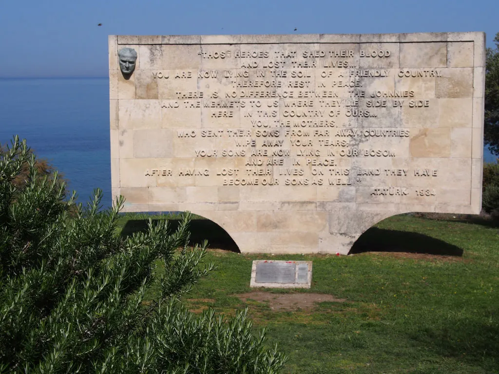 A wide stone memorial plaque stands on a grassy cliff overlooking the blue sea. The monument features a small bust of a man on the top left and a long English inscription beginning with "Those heroes that shed their blood..." attributed to Atatürk, 1934.