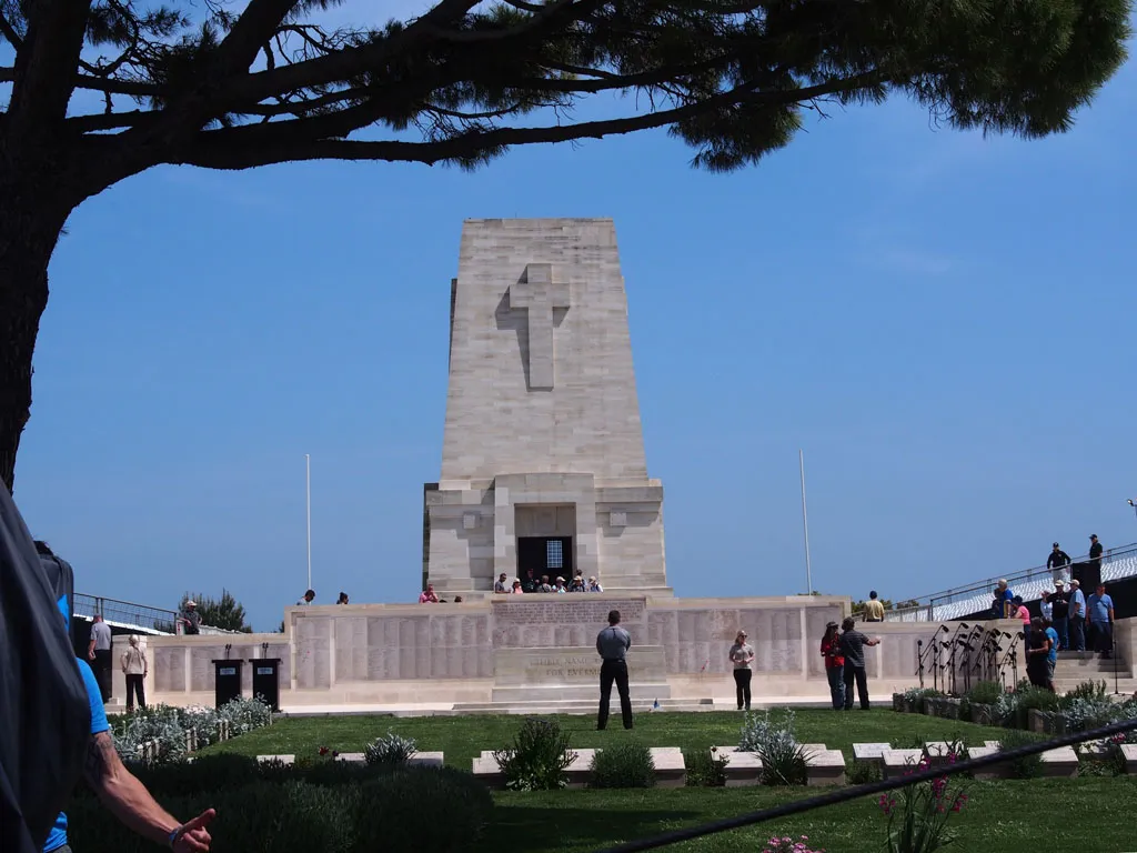 A large, light-colored stone memorial pylon with a carved cross stands under a clear blue sky, surrounded by visitors and uniform grave markers.