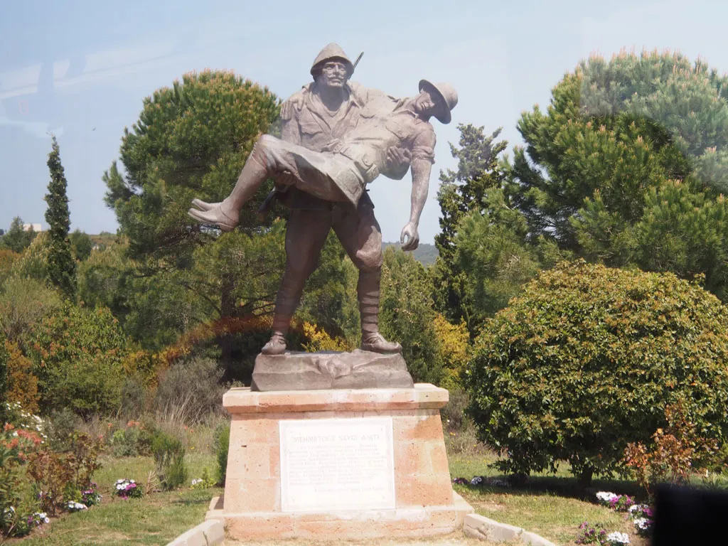 A bronze statue depicting a Turkish soldier heroically carrying a wounded Allied soldier. The statue stands on a stone pedestal with an inscribed plaque, surrounded by green trees and flowering bushes.