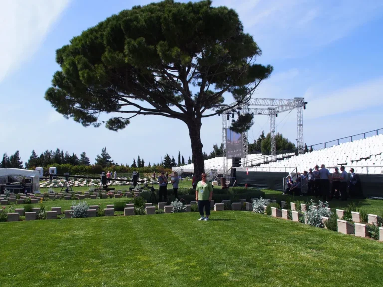 A woman stands on a lush green lawn in a military cemetery, positioned beneath a large, spreading pine tree. In the background, white stadium-style seating and a large stage structure have been erected for a commemorative event.