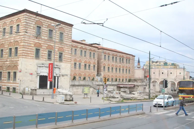 A multi-story historic building with a distinctive red and tan brick pattern stands at a busy intersection with a tram line, a white car, and a passing yellow bus.