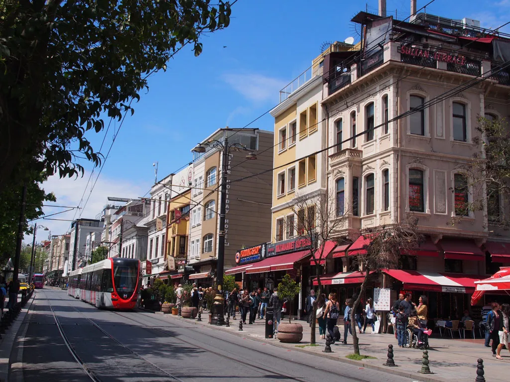 A street-level view of a wide Istanbul city street featuring a modern red and white tram on tracks, historic multi-story buildings with red awnings, and pedestrians on a sunny day.
