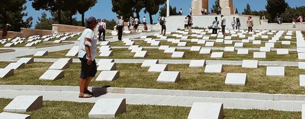 Visitors walk among rows of white headstones on a grassy slope leading up to a large stone memorial cross.