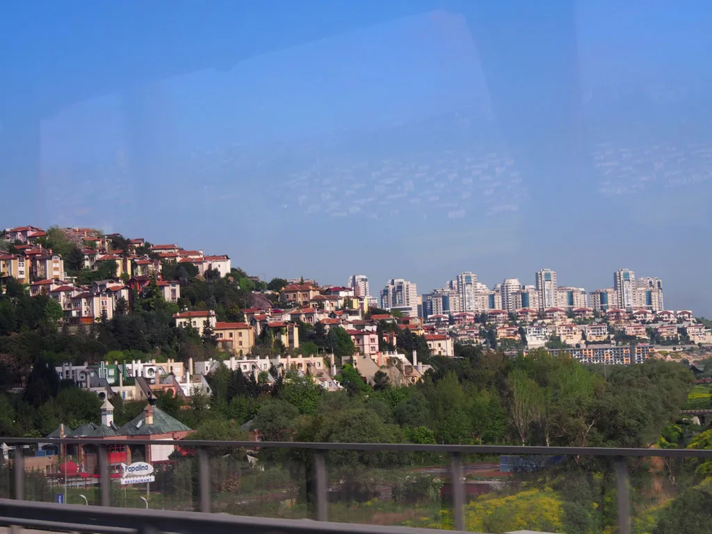 A view from a vehicle of a lush green hillside dotted with traditional houses with red roofs on the left, transitioning to a dense cluster of modern, white high-rise apartment buildings on the right under a clear blue sky.