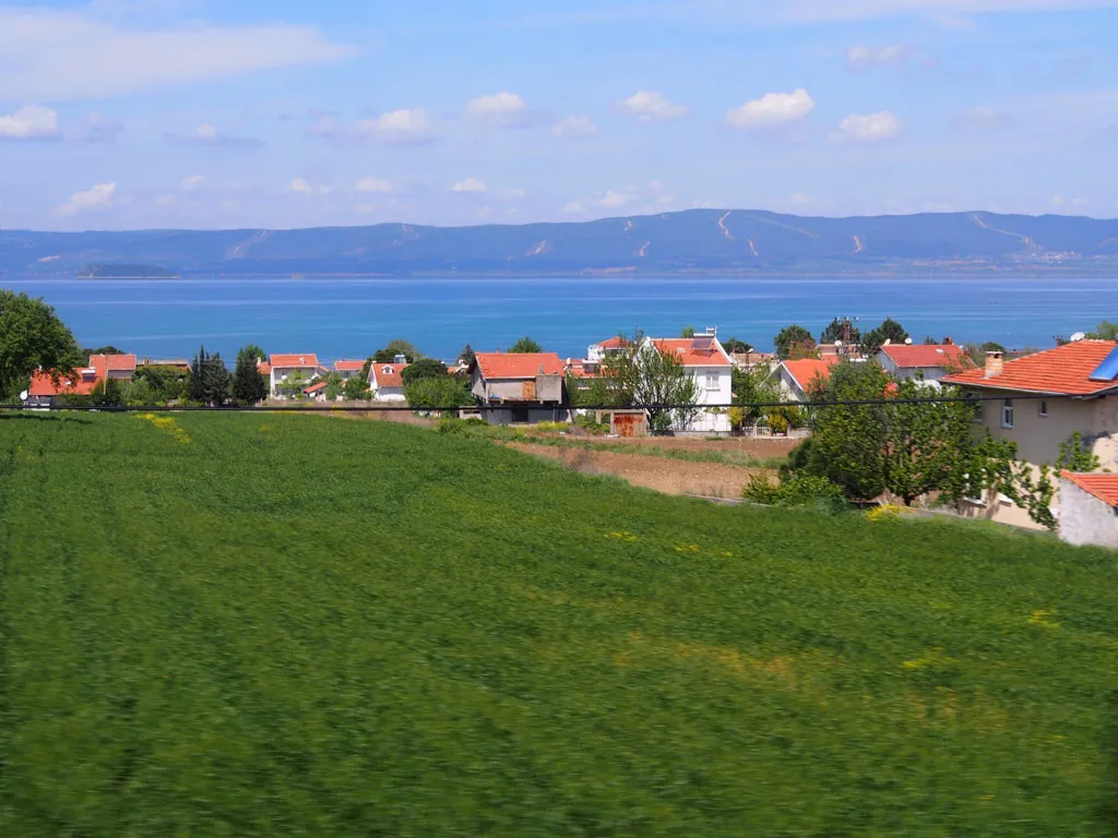 A scenic view of a lush green field in the foreground leading to a small village with orange-roofed houses, situated along a large blue body of water with mountains in the distance.