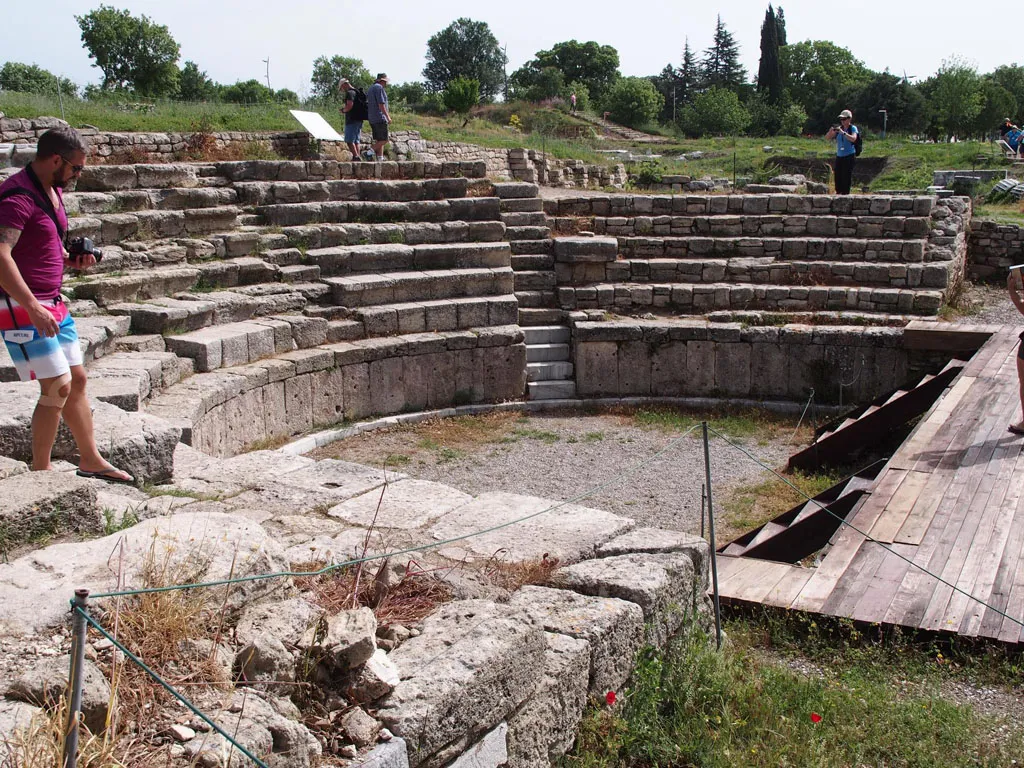 A view of an ancient semi-circular stone theater with tiered seating, featuring a small central stage area and several tourists exploring the ruins under a bright sky.