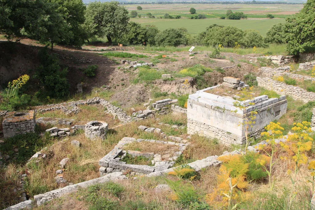 A high-angle view of an archaeological excavation site featuring ancient stone wall foundations, stone-lined wells, and a larger rectangular stone structure nestled in a grassy hillside.