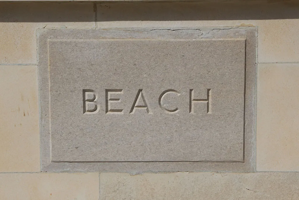 A close-up of a light-colored rectangular stone block with the word "BEACH" carved in simple, serif capital letters.