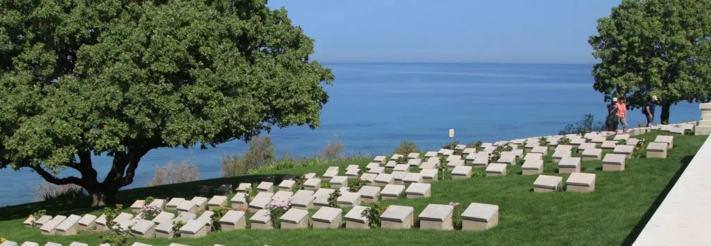 A scenic view of a military cemetery situated on a green grassy slope overlooking a deep blue sea. Neatly arranged rows of uniform, light-colored headstones lead the eye toward the water. On the left, a large, lush green tree provides shade, while a few visitors walk along the perimeter on the right.