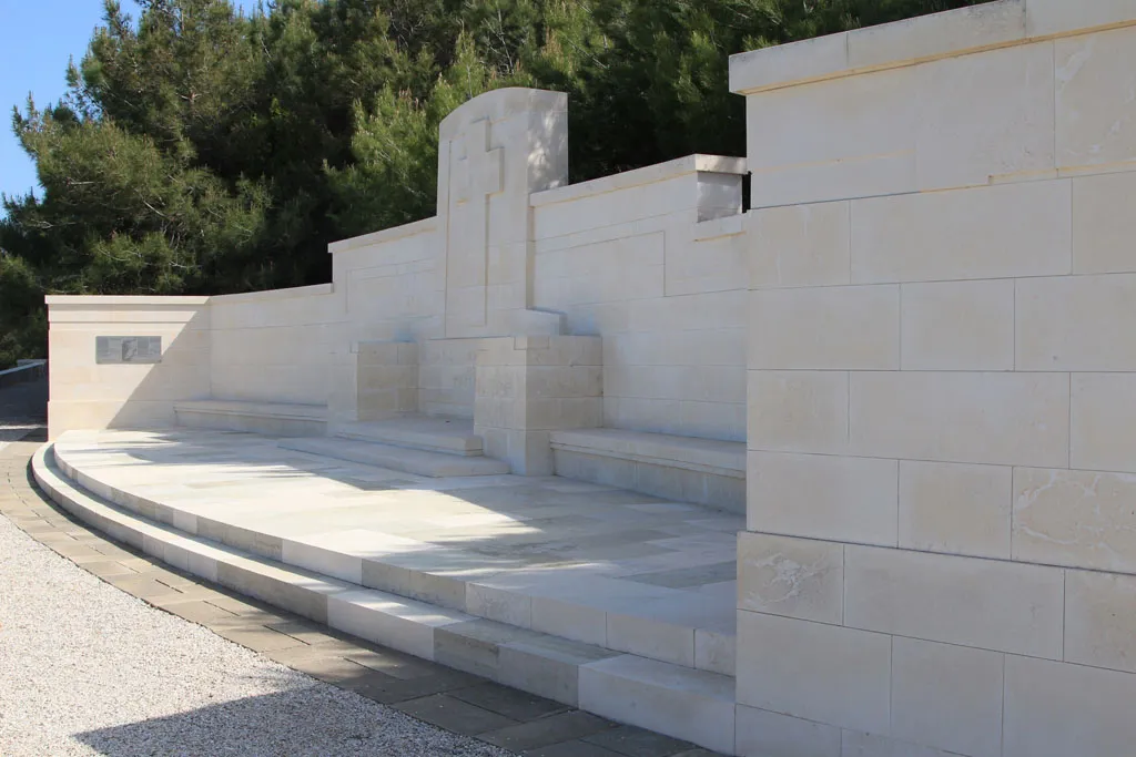 A large, curved memorial wall made of smooth, light-colored stone blocks, featuring a central engraved cross and stone benches under a clear sky.