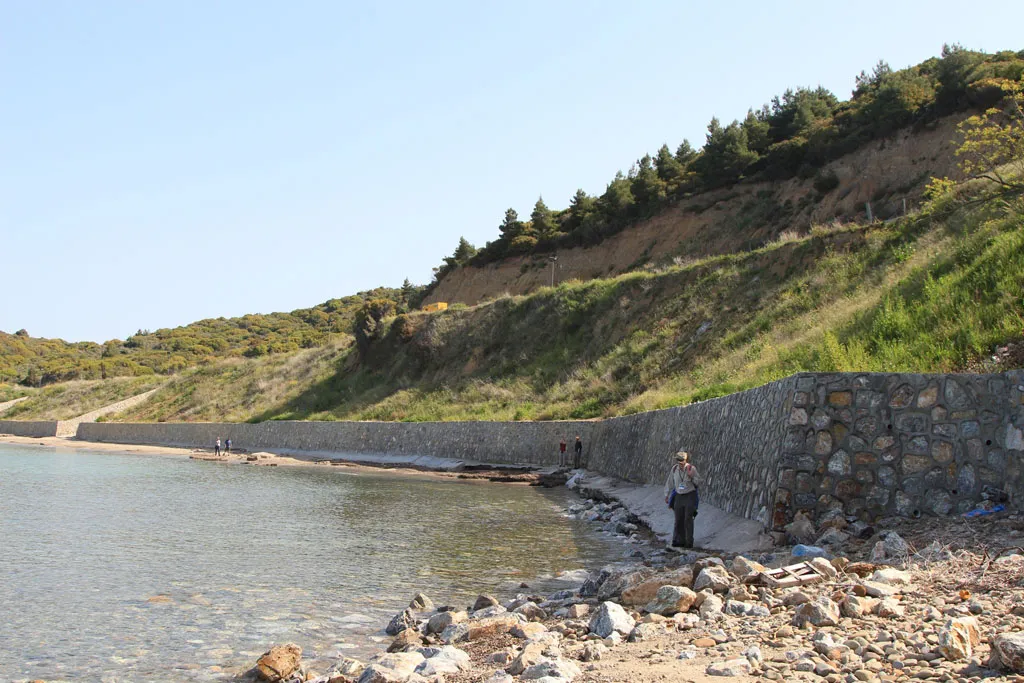 A view of a rocky shoreline featuring a long, sturdy sea wall made of grey stones. The wall follows the curve of the coast beneath a steep, grassy hillside topped with evergreen trees. A few people are visible walking along the narrow beach area near the water's edge under a clear, bright sky.