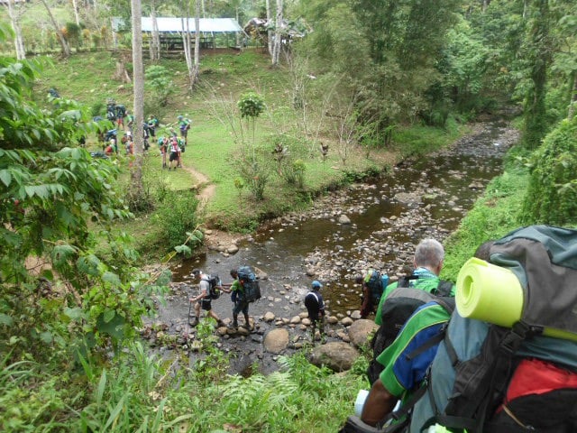 A high-angle view shows a line of trekkers with large backpacks carefully crossing a shallow, rocky stream. They are stepping on large stones to keep their feet dry. On the far bank, a grassy clearing leads toward a building with a blue metal roof, where other team members are gathered.