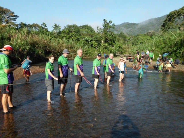 A group of about ten trekkers in matching green and purple jerseys are wading through a shallow, wide river. They are holding onto a safety rope stretched across the water for balance. The riverbank is lush with green tropical trees and tall grass under a clear blue sky.