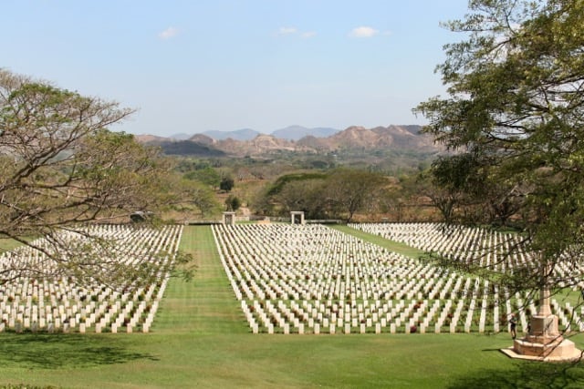 Expansive military cemetery with rows of white headstones arranged symmetrically across a green lawn, surrounded by trees and hills, with stone memorial structures visible under a clear sky.