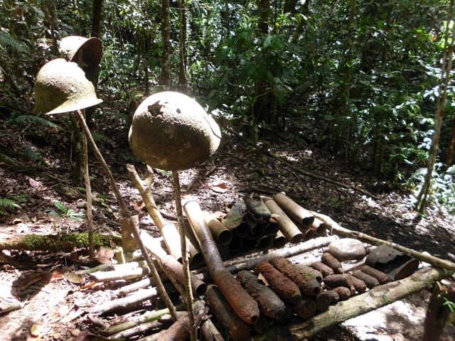 In a dense, sun-dappled forest, several rusted and pitted military helmets are perched on top of upright wooden sticks. Below them, a makeshift wooden platform holds a collection of corroded artillery shells and mortar rounds of various sizes. The scene is quiet and overgrown, serving as a primitive memorial to past conflict.