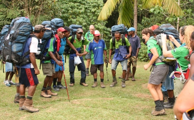 A diverse group of hikers and local guides wearing large backpacks gather in a grassy clearing, talking and laughing together.