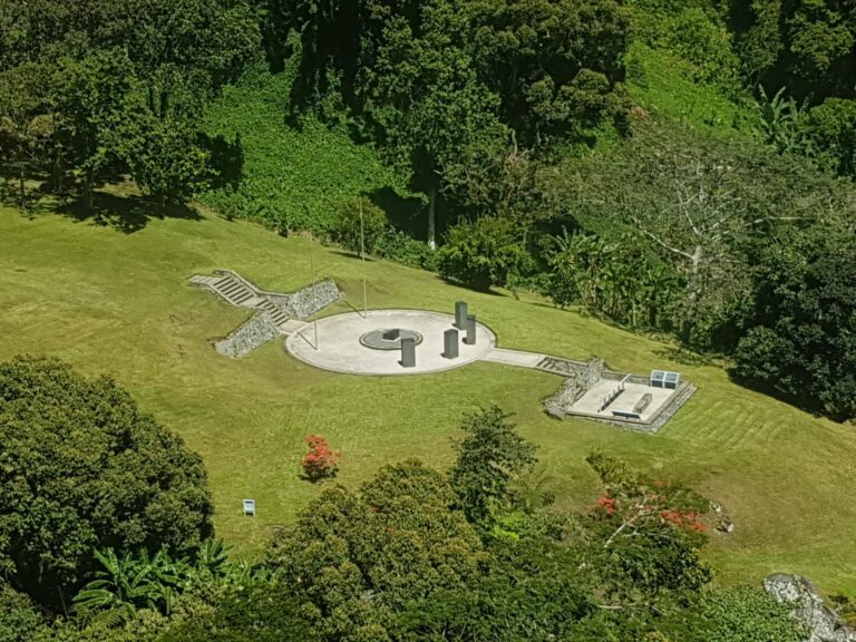 An aerial view of a stone and concrete memorial site nestled in a lush green clearing surrounded by dense forest.