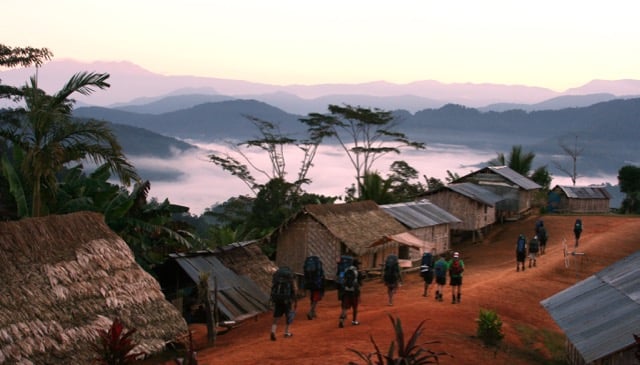 A group of hikers with large backpacks walk along a dirt path through a remote mountain village at dawn, overlooking a valley filled with thick mist.