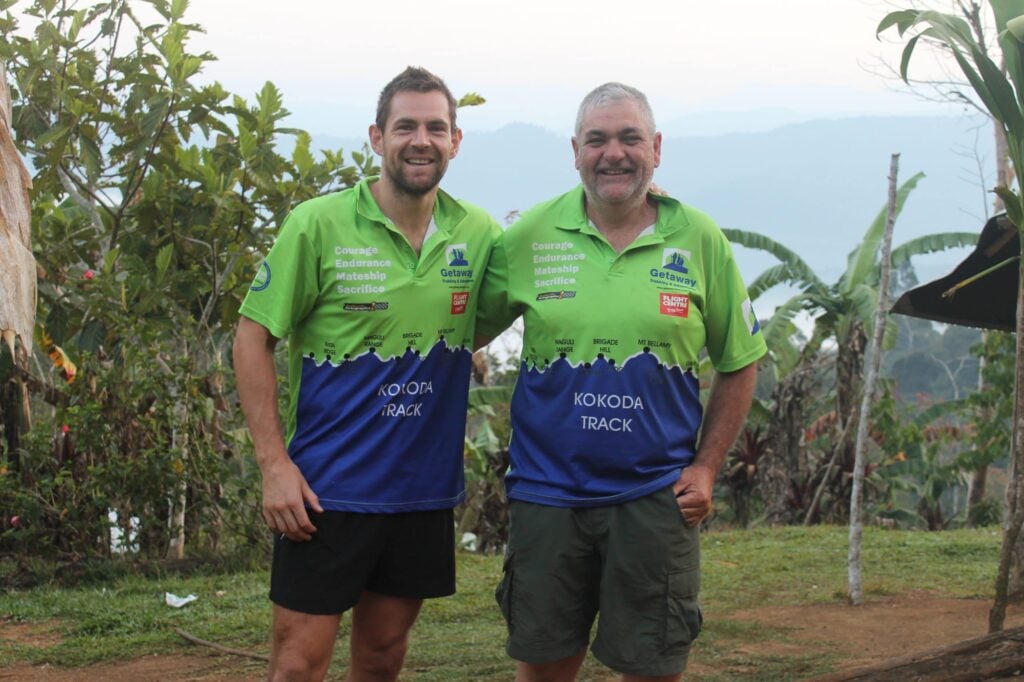 Two men pose together outdoors wearing matching green and blue athletic shirts that feature "Kokoda Track" and the words "Courage, Endurance, Mateship, Sacrifice".