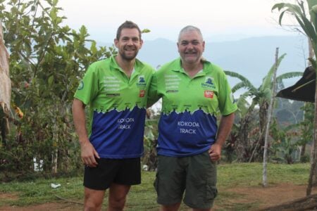 Two men pose together outdoors wearing matching green and blue athletic shirts that feature "Kokoda Track" and the words "Courage, Endurance, Mateship, Sacrifice".