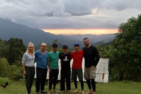 Six people in athletic wear stand together in a grassy field in front of a corrugated metal building. A rugby ball sits on the ground in front of them, and a dramatic sunset glows behind a deep mountain range in the background.
